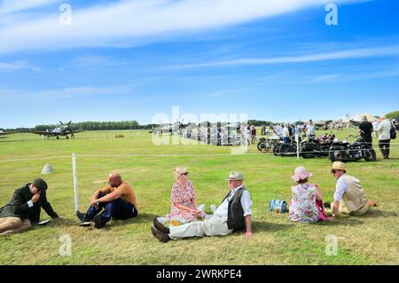 Persone sedute a rilassarsi sul campo d'aviazione in erba soleggiata vestite con abiti retrò al Goodwood Revival con motociclette classiche e aerei d'epoca, Regno Unito Foto Stock