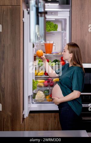 Giovane donna incinta con un'arancia vicino al frigorifero di casa. Mangiare sano. Foto Stock