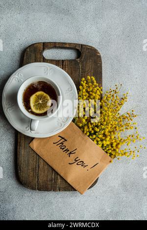 Una vista dall'alto di una classica tazza da tè con limone, posta su una tavola di legno, accompagnata da luminosi fiori di Mimosa e un grazie! scheda su un dorso testurizzato Foto Stock
