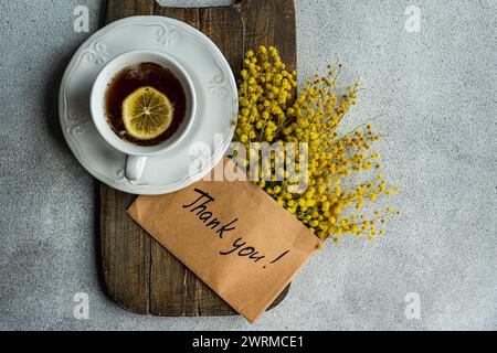 Una vista dall'alto di una classica tazza da tè con limone, posta su una tavola di legno, accompagnata da luminosi fiori di Mimosa e un grazie! scheda su un dorso testurizzato Foto Stock