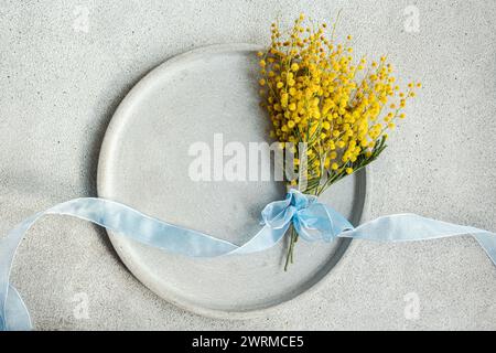 Vista dall'alto del pittoresco bouquet di fiori di mimosa, delicatamente legato con un nastro azzurro, poggia su un piatto di ceramica con uno sfondo di cemento liscio Foto Stock