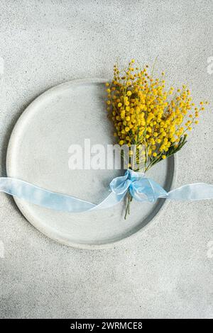 Vista dall'alto del pittoresco bouquet di fiori di mimosa, delicatamente legato con un nastro azzurro, poggia su un piatto di ceramica con uno sfondo di cemento liscio Foto Stock