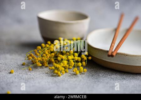 Un ambiente tranquillo con stoviglie in ceramica con bacchette di rame e una dispersione di luminosi fiori di mimosa che aggiungono un tocco di colore Foto Stock