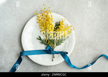 Dall'alto del piatto bianco ornato contiene un mazzo luminoso di fiori di mimosa, elegantemente legato con un nastro blu profondo su una superficie grigia testurizzata Foto Stock