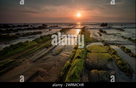 Maestoso tramonto sulle rocce coperte di muschio a Barrika Beach in Biscaglia, Spagna, che proietta un caldo bagliore sulla costa frastagliata. Foto Stock