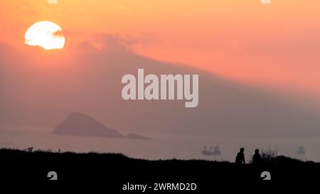 Un tranquillo paesaggio marino con la silhouette di una coppia contro un morbido tramonto arancione, accanto a navi lontane e a una montagna, il luogo è Cabo Peñas, Asturie Foto Stock