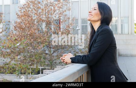 Una donna professionista con un costume da lavoro è in piedi fuori, appoggiata su una ringhiera e sorridente mentre si gode una pausa tranquilla. Foto Stock