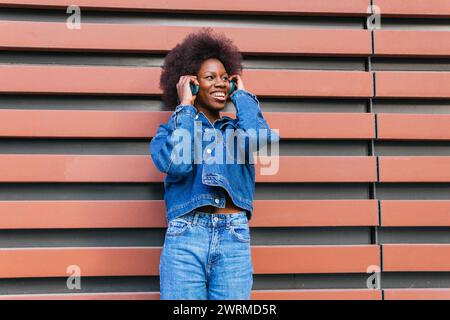 Una donna radiosa ama le sue canzoni preferite, vestita testa a piedi con jeans classici su uno sfondo geometrico in terracotta Foto Stock