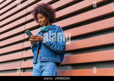 Dal basso di una giovane donna allegra con le cuffie intorno al collo sorride mentre utilizza il suo smartphone, appoggiandosi su una parete a righe arancioni Foto Stock