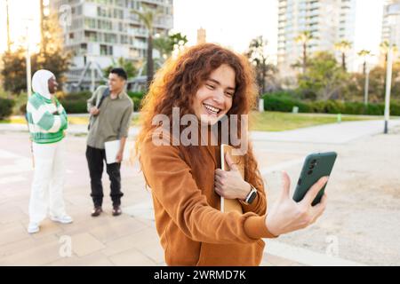 Una donna gioiosa che utilizza uno smartphone con diversi studenti che chiacchierano sullo sfondo in un parco, evidenziando l'interconnettività e l'amicizia. Foto Stock