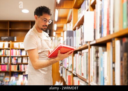 Un giovane con gli occhiali legge intensamente un libro mentre si trova in una sala della biblioteca, circondato da scaffali pieni di titoli diversi. Foto Stock