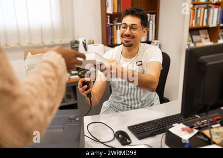 Un allegro cassiere maschile in grembiule a righe che scansiona gli articoli al momento della cassa di un negozio mentre sorride al cliente. Foto Stock