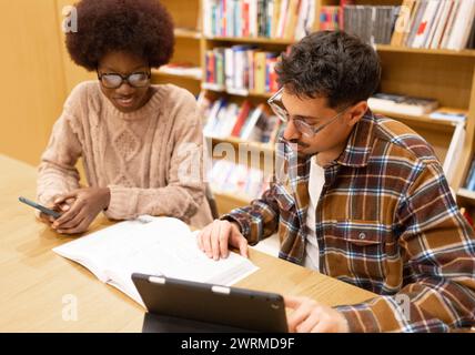 Una giovane donna afroamericana e un uomo siedono a un tavolo in una biblioteca, concentrati sul loro lavoro la donna afroamericana usa un telefono cellulare mentre la m Foto Stock
