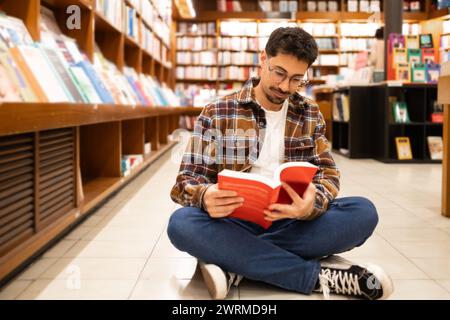 Un giovane uomo siede a gambe incrociate sul pavimento di una libreria, avvolto da un libro rosso, circondato da scaffali pieni di libri. Foto Stock