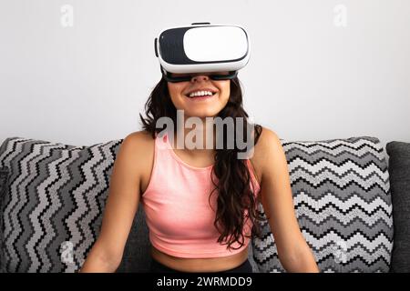 Una giovane e allegra donna ispanica adulta indossa un visore per la realtà virtuale seduto sul divano di casa Foto Stock