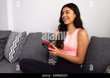 Una donna allegra si siede comodamente su un divano, impegnata a giocare a un videogame con un controller wireless. Foto Stock