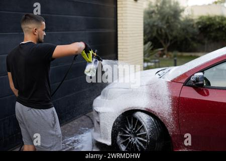 Un giovane uomo viene mostrato con un'irroratrice ad alta pressione per lavare un'auto rossa, concentrandosi sul processo di pulizia Foto Stock