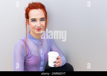 Una giovane donna d'affari trucco blu che mette in risalto gli occhi tenendo in mano una tazza di caffè da asporto bianca con una camicetta lilla trasparente che guarda la fotocamera Foto Stock