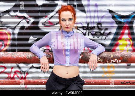Una donna caucasica si posa su uno sfondo spigoloso di graffiti colorati. I suoi sorprendenti capelli rossi contrastano con la sua blusa lilla trasparente e i pantaloni neri Foto Stock