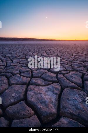 Le sfumature radiose di un sole che tramonta illuminano gli intricati schemi di profonde crepe di fango, creando un contrasto tra terra arida e un cielo vibrante Foto Stock