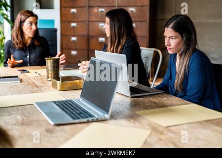 Tre colleghi si impegnano in una discussione a un tavolo di legno con computer portatili, esprimendo il lavoro di squadra all'interno di un'azienda guidata da donne. Foto Stock