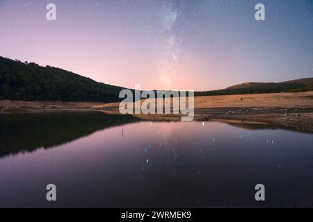 Un lago sereno riflette uno splendido cielo stellato notturno con la via Lattea, circondato da un paesaggio aspro con crepe fangose visibili, che evocano un senso di pisello Foto Stock