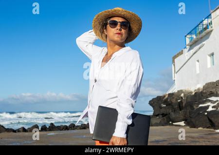 Una nomade digitale si trova in un villaggio di pescatori estivo a Fuerteventura, con un portatile in mano, pronto a lavorare con l'oceano come un backd Foto Stock