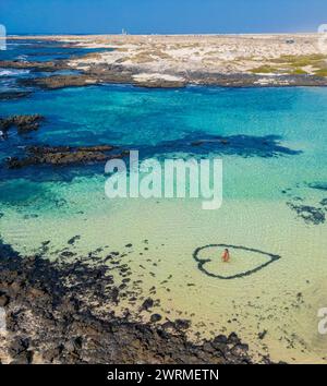 Una vista aerea cattura una donna in piedi all'interno di una formazione naturale a forma di cuore nelle acque cristalline della costa, con terreno vulcanico nel bac Foto Stock