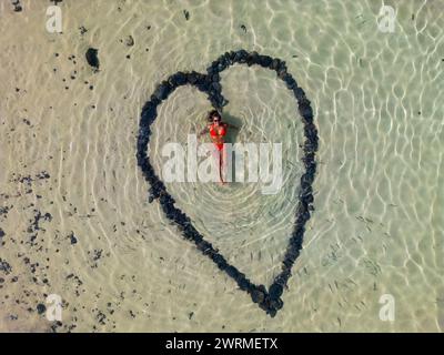 Un'immagine aerea cattura una donna sdraiata all'interno di una forma di cuore formata da pietre su una spiaggia tranquilla con acque cristalline che brillano al tramonto. Foto Stock