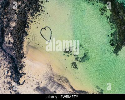 Vista aerea di un segno naturale a forma di cuore sulla spiaggia con acqua cristallina vicino al vulcano Calderon Hondo a Fuerteventura durante il tramonto. Foto Stock