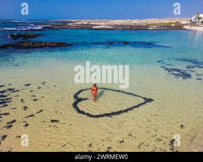 Vista aerea di una donna che si trova all'interno di un cuore disegnato sul fondo sabbioso di una spiaggia di acqua cristallina, con il vulcano Calderon Hondo visto al tramonto Foto Stock