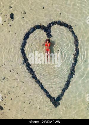 Vista aerea di una formazione a forma di cuore fatta di pietre su una spiaggia sabbiosa con una donna sdraiata al centro, catturando l'essenza dell'amore estivo. Foto Stock