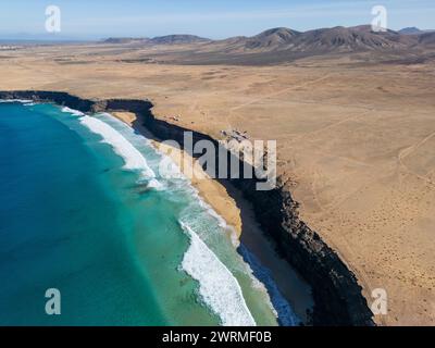 Immagine aerea di un drone che cattura il contrasto mozzafiato tra l'azzurro oceano e l'arido paesaggio della costa frastagliata di Fuerteventura. Foto Stock