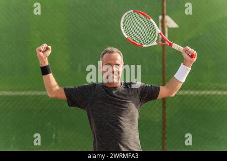 Un tennista maschio gioioso celebra una vittoria sul campo da tennis, reggendo trionfalmente la sua racchetta e guardando la telecamera con un sorriso orgoglioso. Foto Stock