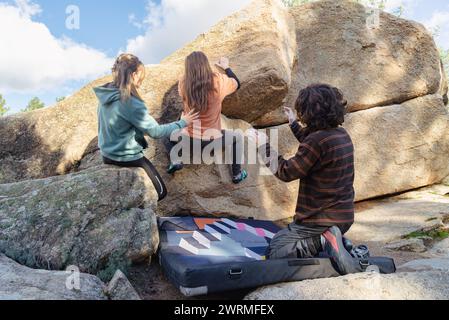 La vista posteriore di tre amici si impegna a boulder su una grande roccia, con un solo avvistamento per sicurezza, su uno sfondo di cieli limpidi Foto Stock