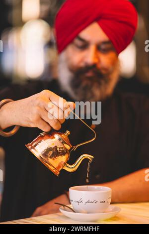 Un uomo con un turbante rosso che versa con cura il tè da una teiera di rame in una tazza bianca su un tavolo di legno Foto Stock