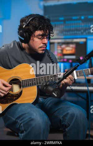 Un musicista concentrato che suona una chitarra acustica durante una sessione in studio, con apparecchiature di registrazione in sottofondo. Foto Stock