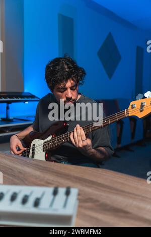 Un musicista maschile attento suona sapientemente un basso durante una sessione di registrazione in studio, con attrezzature musicali in sottofondo. Foto Stock