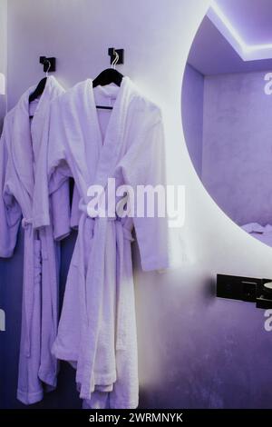 Due accappatoi bianchi su grucce. Interno del salone di bellezza. Accappatoi di cotone nel centro benessere. Abiti accoglienti in camera da letto. Resort di lusso al coperto. Foto Stock