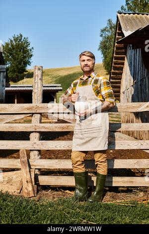 uomo dall'aspetto gradevole con tatuaggi sulle braccia che reggono il vaso di latte fresco e guardano la macchina fotografica mentre si è in fattoria Foto Stock