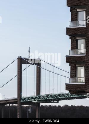 Il lato di un edificio residenziale in mattoni con i suoi moderni balconi si accostano alle linee architettoniche di un lontano ponte sospeso verde nel Get Foto Stock