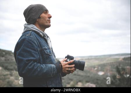 Ritratto laterale sicuro di un giovane uomo attraente, un fotografo di viaggio che tiene in mano una fotocamera digitale professionale, che cattura una splendida vista mentre cammina in m Foto Stock