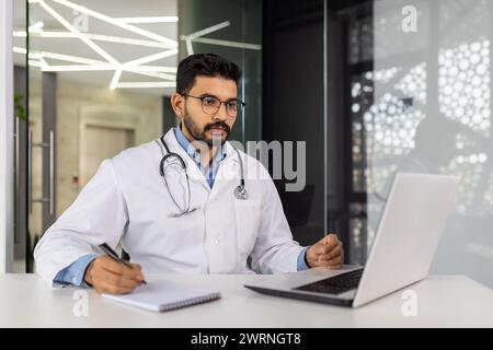 Medico maschile concentrato con stetoscopio presso la stazione di lavoro clinica, scrivendo appunti mentre si guarda lo schermo del computer portatile. Foto Stock