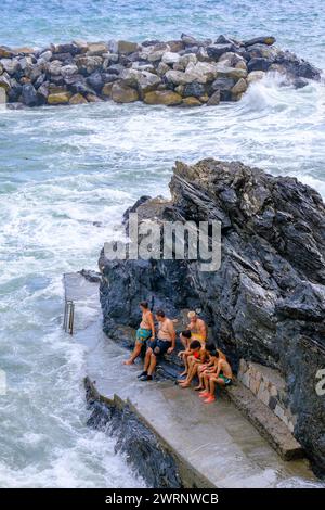 Manarola, IT - 25 luglio 2023: I giovani rimangono su una roccia mentre grandi onde si infrangono sulla costa Foto Stock