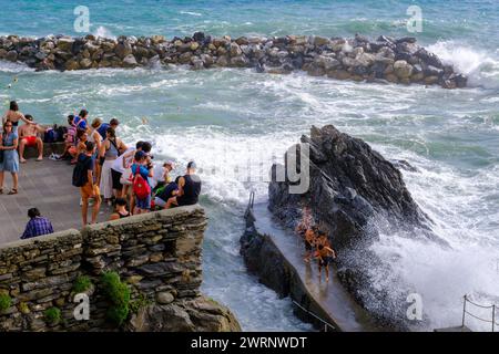 Manarola, IT - 25 luglio 2023: I giovani rimangono su una roccia mentre grandi onde si infrangono sulla costa Foto Stock