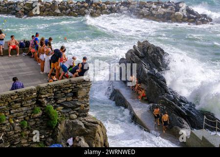 Manarola, IT - 25 luglio 2023: I giovani rimangono su una roccia mentre grandi onde si infrangono sulla costa Foto Stock