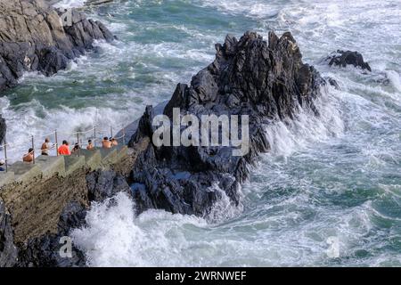 Manarola, IT - 25 luglio 2023: I giovani rimangono su una roccia mentre grandi onde si infrangono sulla costa Foto Stock