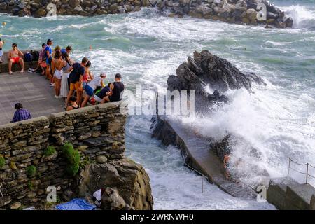 Manarola, IT - 25 luglio 2023: I giovani rimangono su una roccia mentre grandi onde si infrangono sulla costa Foto Stock