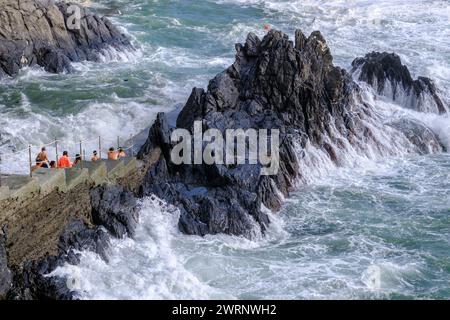 Manarola, IT - 25 luglio 2023: I giovani rimangono su una roccia mentre grandi onde si infrangono sulla costa Foto Stock
