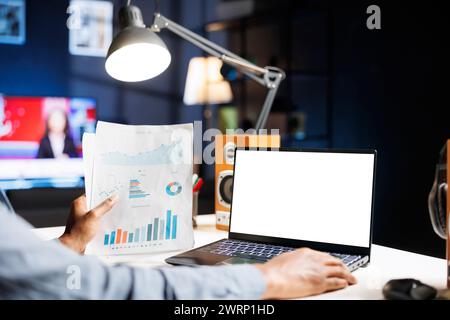 L'analista afroamericano esamina i file su schermo bianco e report , lavorando in remoto da casa e utilizzando un display isolato di copyspace su un computer portatile. L'esperto esamina modelli e documenti vuoti. Foto Stock